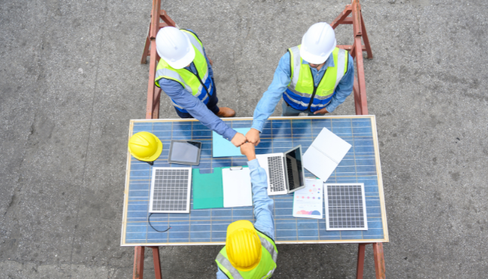 Three engineers wearing safety vests and helmets shake hands over a table covered with solar panels and documents, symbolizing teamwork in Electricity in the US projects.