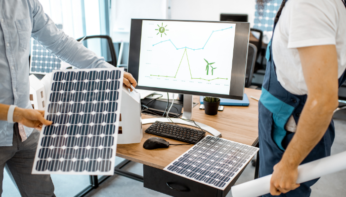 Two professionals hold solar panels while reviewing energy data on a computer screen, illustrating the planning and innovation