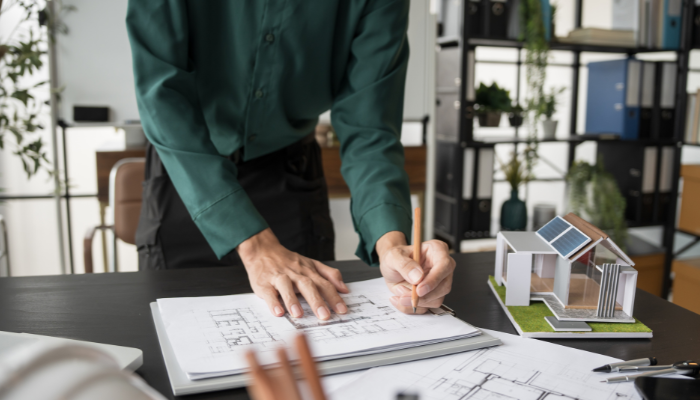 A person sketches building plans on a desk with a scale model of a solar-powered house, illustrating the planning and design of Energy Projects in US.