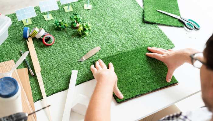 Un profesional manipula césped artificial sobre una mesa de trabajo con miniaturas de paneles solares y árboles, ilustrando la implementación de Medidores inteligentes en España y la apuesta por soluciones sostenibles.