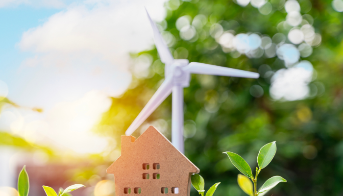 Una maqueta de casa recortada en cartón se sitúa frente a una turbina eólica difuminada, representando la importancia de las energías limpias en el Mercado Energético en Argentina.