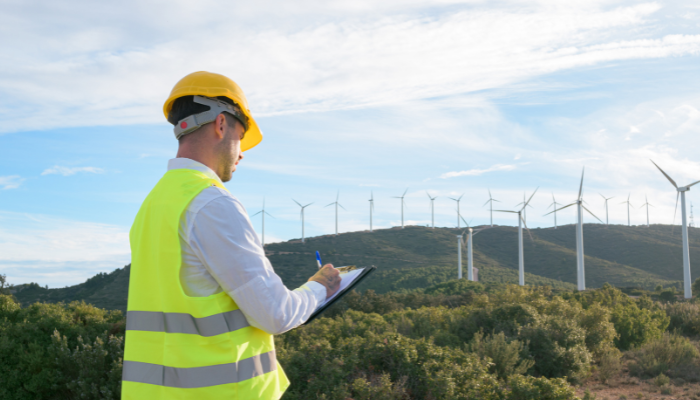 Un profesional con chaleco reflectante y casco toma notas en un bloc, mientras se observan aerogeneradores en una colina al fondo, ilustrando la supervisión del Mercado Energético en España