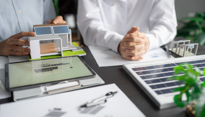Dos especialistas revisan una maqueta de vivienda con paneles solares sobre una tablet, representando el análisis de proyectos y estrategias