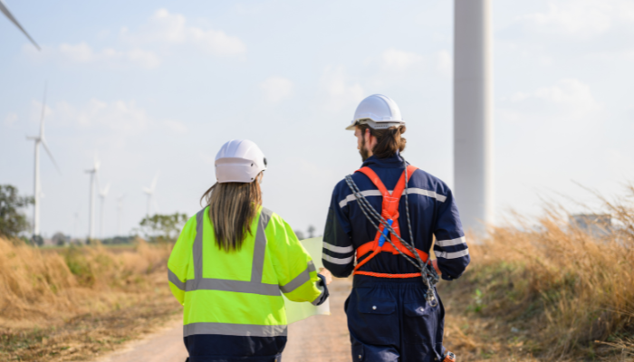 Dos profesionales con cascos y equipo de seguridad caminan por un sendero rodeado de aerogeneradores, ilustrando el trabajo en los Parques Eólicos en España.