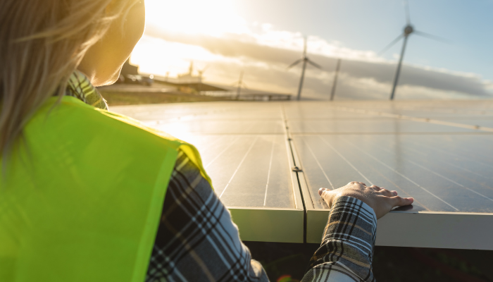 Una ingeniera con chaleco reflectante observa un panel solar mientras se aprecian turbinas de fondo, evidenciando la combinación de fuentes renovables