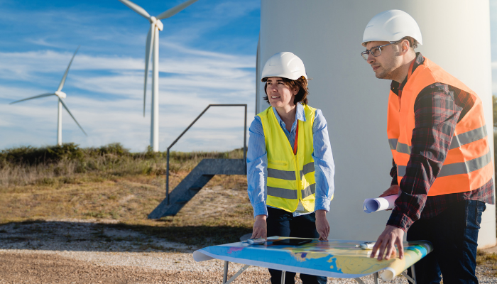 Two engineers wearing safety vests stand by a wind turbine with a map, planning renewable energy infrastructure.