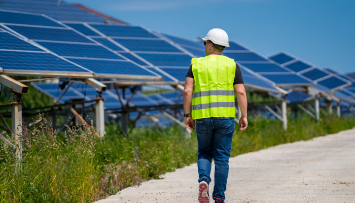 A worker in a reflective vest and helmet walks along a solar farm, highlighting the advancements in Sustainability in the US through renewable energy.