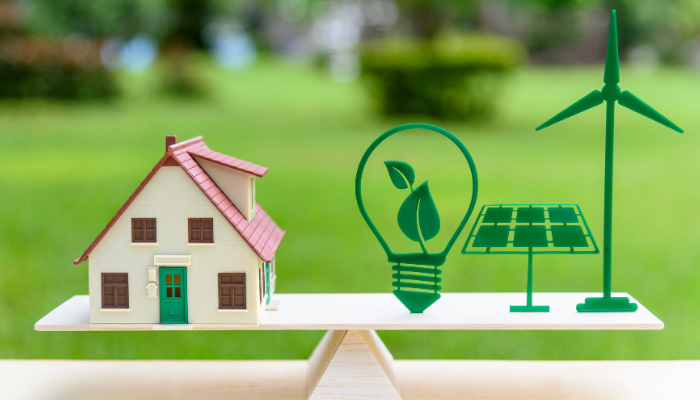 A small house model balances on a wooden plank alongside green icons for a lightbulb, solar panel, and wind turbine, highlighting clean energy initiatives that complement Wind Farms in US.