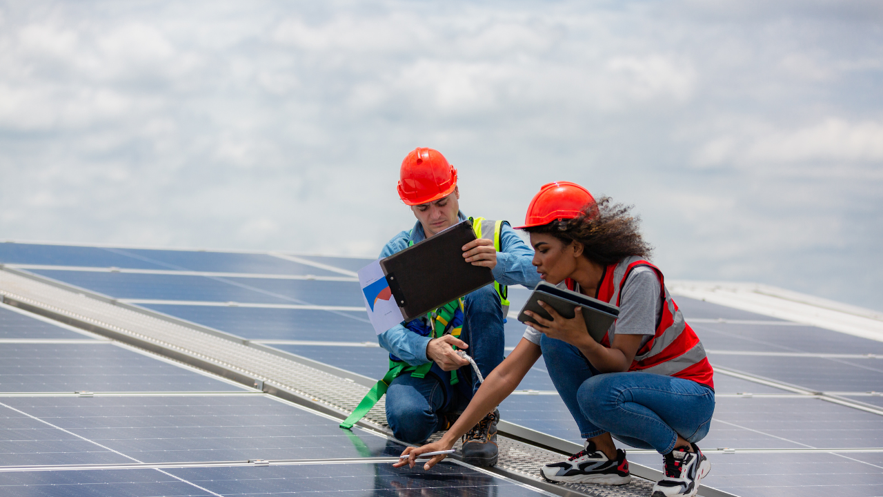 Two engineers, equipped with helmets and safety vests, inspect solar panels using tablets and documents. The image reflects the UK’s professional approach to solar energy for businesses, emphasizing precision and technical oversight.