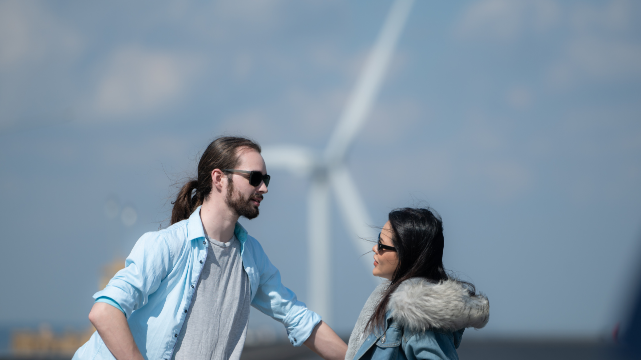 A young couple chats casually outdoors with a large wind turbine towering behind them, suggesting a modern and eco-conscious setting.