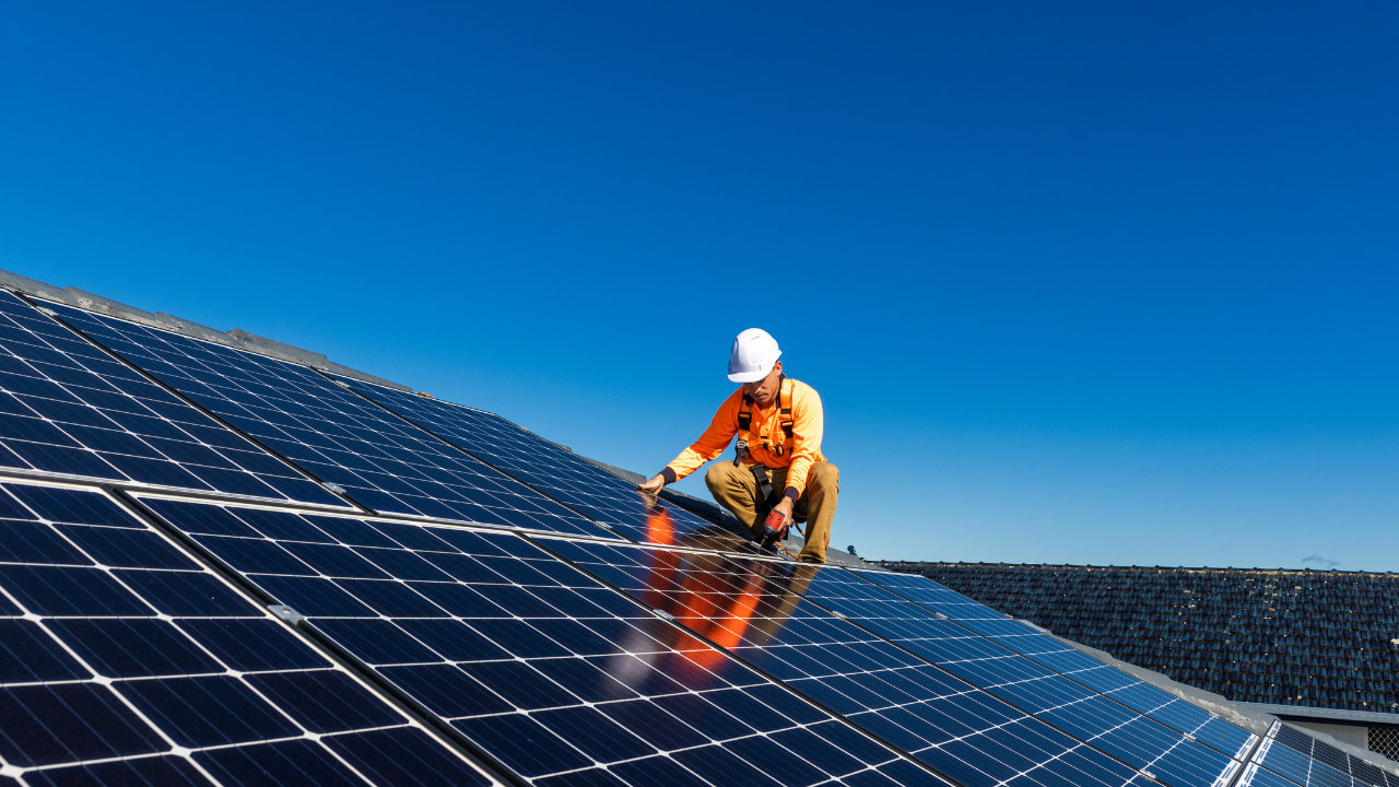 Un técnico con casco blanco y arnés de seguridad instala paneles solares en el techo de una casa bajo un cielo despejado. La escena transmite el crecimiento del uso de paneles solares usados en viviendas argentinas, promoviendo soluciones energéticas más accesibles y sostenibles.