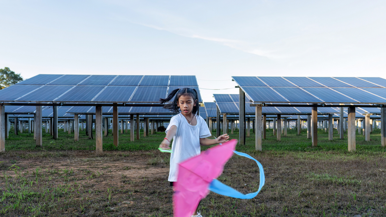 Una niña juega con una cometa de colores brillantes frente a un parque solar, transmitiendo cómo la energía solar se integra de forma natural en el entorno y la vida diaria.