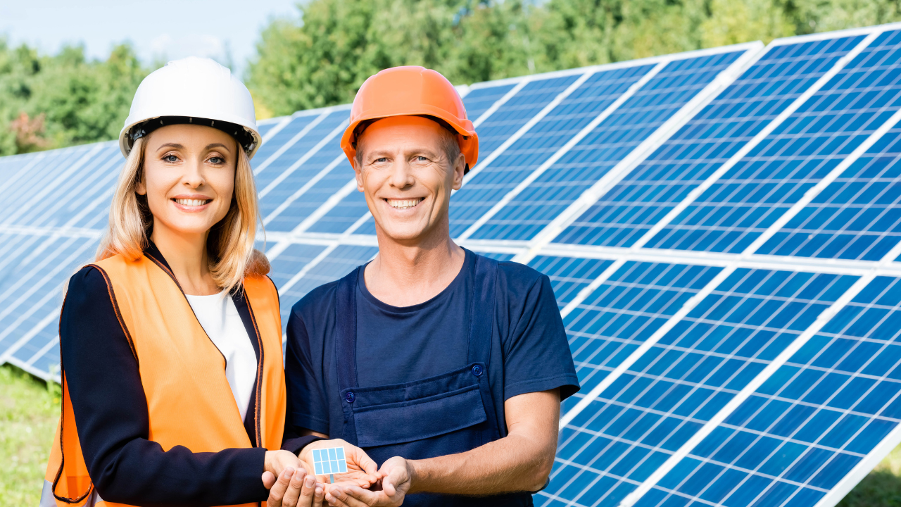 Two engineers, one male and one female, smile while standing in front of a large solar panel array. They proudly hold a miniature model of a solar panel in their hands, embodying the spirit of solar energy innovation and teamwork in a clean energy transition.