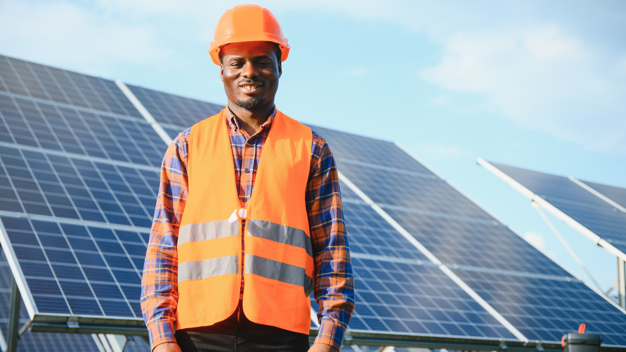 A man in an orange safety vest and helmet stands confidently in front of solar panels under a blue sky. His pose and attire emphasize professional involvement and field expertise, aligned with the pursuit of solar energy development.