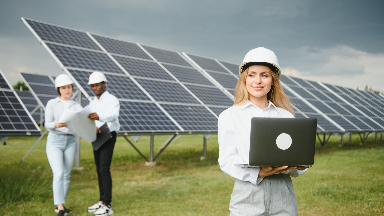 Una ingeniera con casco blanco sostiene una laptop frente a una gran instalación de paneles solares, mientras otros dos colegas revisan planos al fondo. La escena refleja un entorno profesional dedicado al análisis y aprovechamiento de la energía solar.