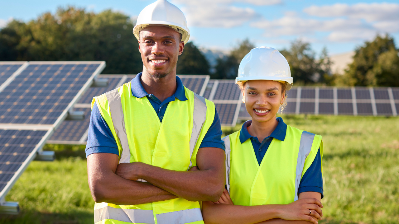 Dos jóvenes ingenieros, un hombre y una mujer, posan sonrientes con chalecos reflectantes frente a un campo de paneles solares. La imagen transmite colaboración y entusiasmo por el trabajo en proyectos de energía solar.