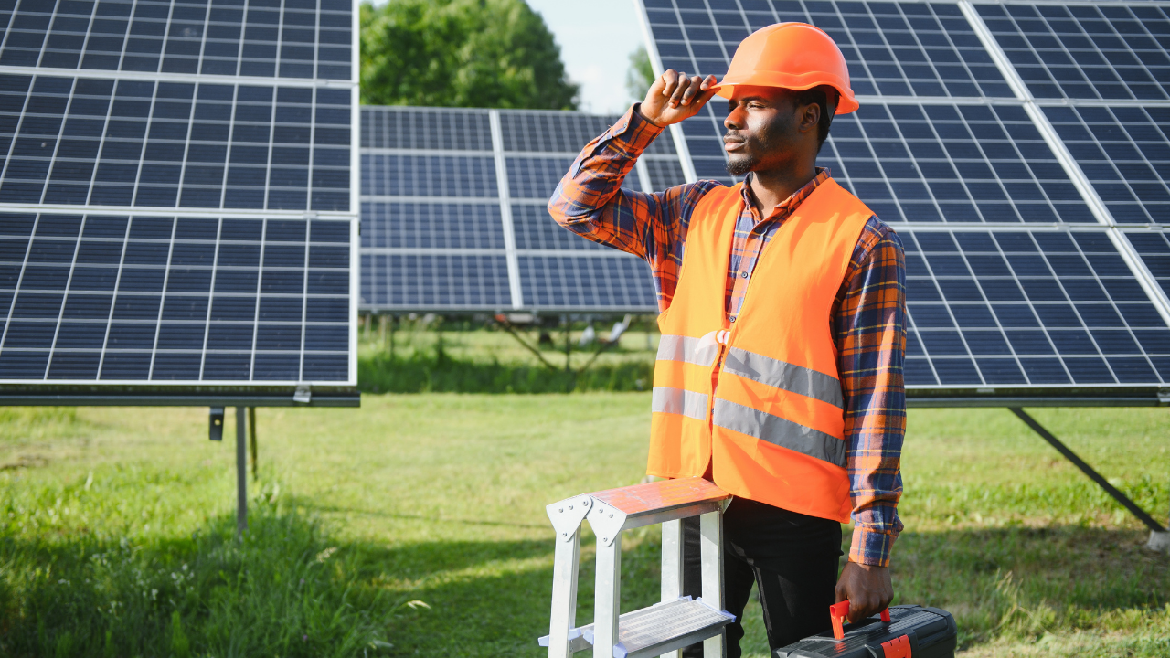 Un técnico con casco naranja y chaleco reflectante observa el horizonte junto a una escalera portátil, rodeado de paneles solares. La escena destaca el compromiso operativo y de mantenimiento necesario para garantizar una producción eficiente de energía solar.