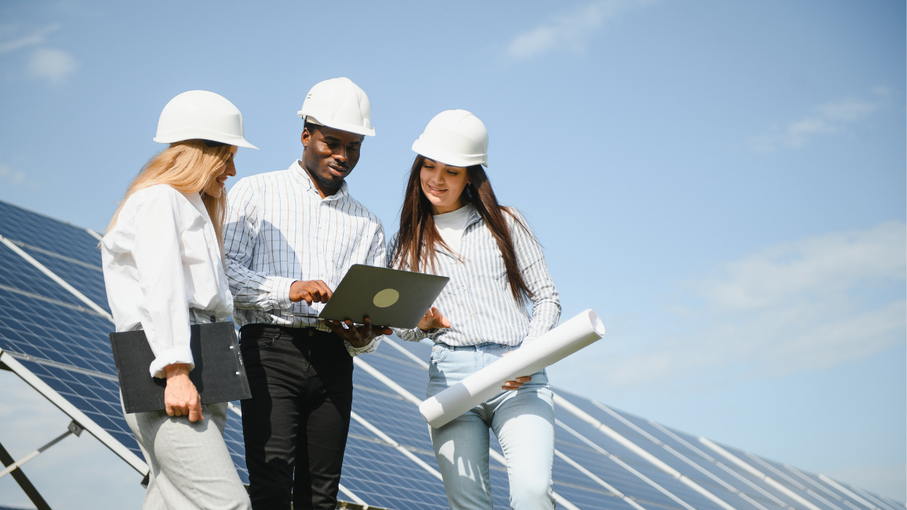 Dos mujeres y un hombre revisan planos junto a paneles solares bajo un cielo despejado. La interacción colaborativa y el uso de tecnología representan claramente el concepto de autoconsumo compartido aplicado en proyectos energéticos conjuntos.