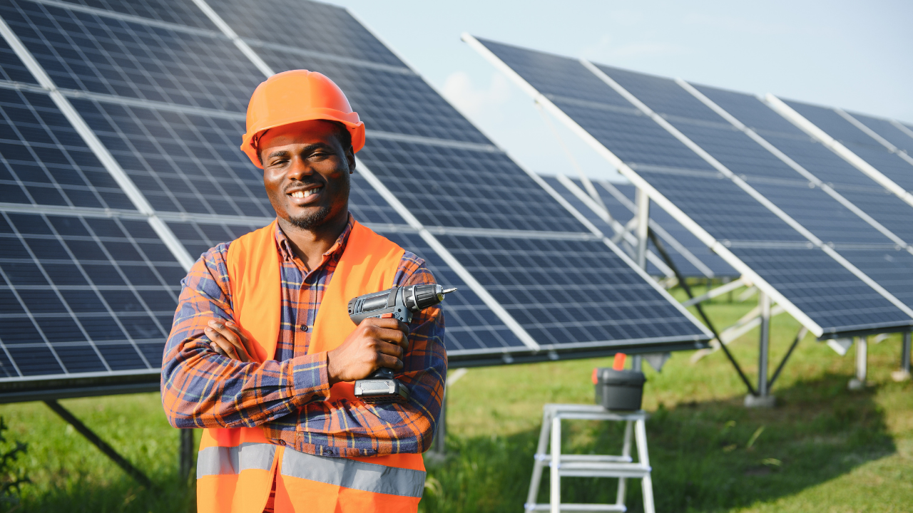 Un trabajador con casco naranja y chaleco reflectante sostiene un taladro con una expresión de confianza frente a una hilera de paneles solares. La escena sugiere una contribución individual en un sistema de autoconsumo compartido.