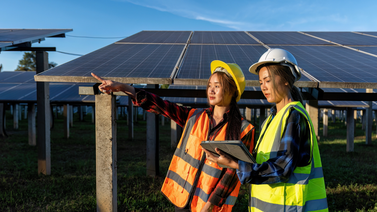 Under a clear blue sky, two women in safety gear stand beside solar panels, one pointing while the other reviews data on a tablet. The image conveys a sense of precision, innovation, and environmental responsibility.
