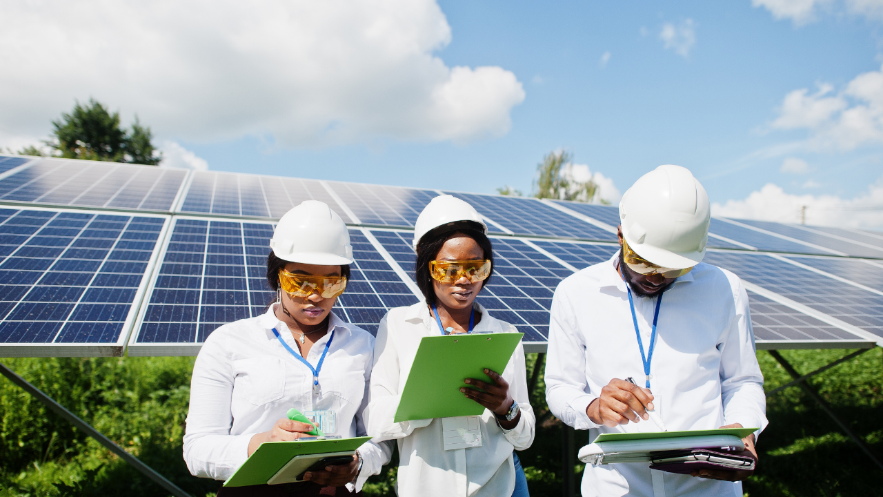 Tres técnicos, equipados con cascos y gafas protectoras, revisan documentos frente a una instalación de paneles solares. Están concentrados en su tarea, representando profesionalismo y compromiso con la transición energética en España.