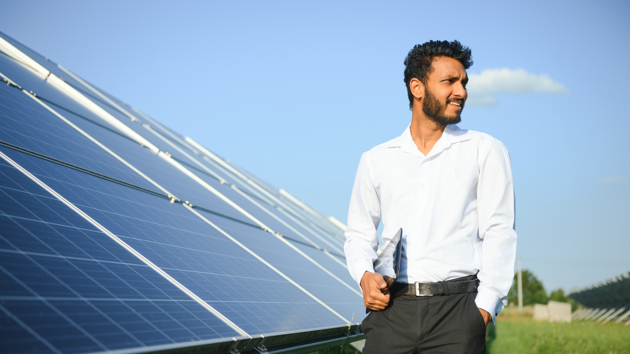 A man in a white shirt stands confidently beside solar panels, holding a tablet and observing the field. His stance and attire reflect a clean, professional approach typical of UK standards to install solar panels.