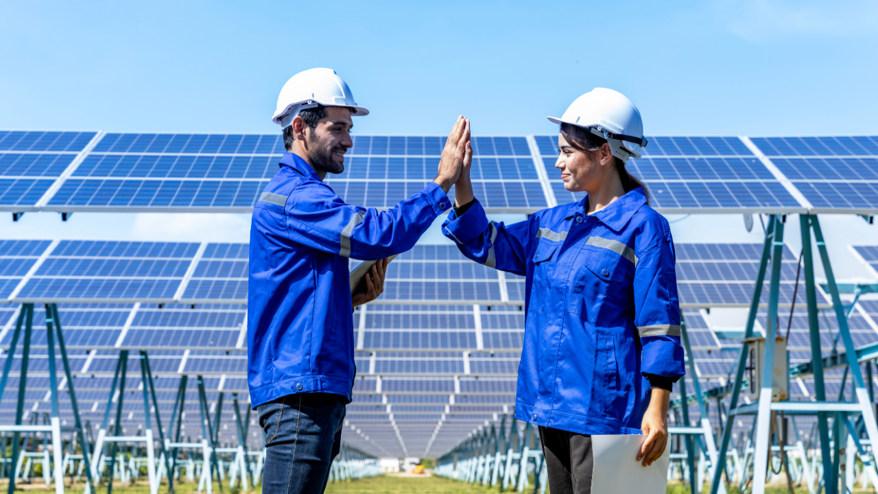Two solar technicians in blue uniforms and white helmets exchange a high five in front of a vast array of solar panels. The cheerful moment conveys teamwork and innovation in renewable energy through portable solar panels.