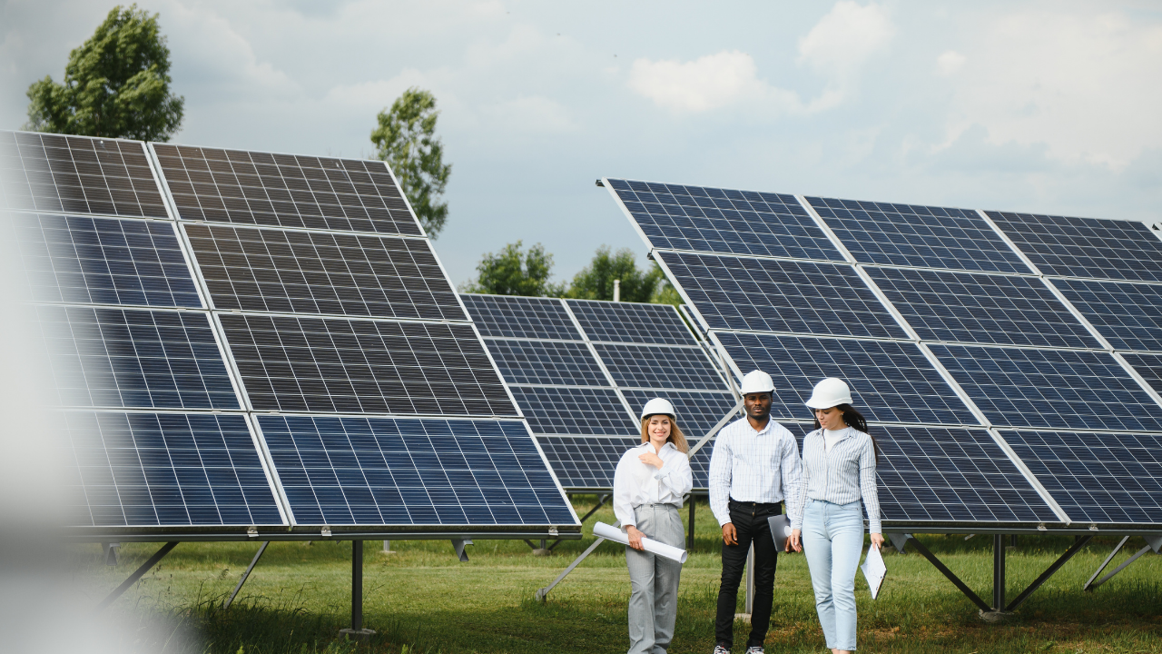 Three professionals in hard hats walk through a solar panel field, appearing to discuss planning or funding opportunities. The context suggests a review of site development possibly supported by grants and subsidies.
