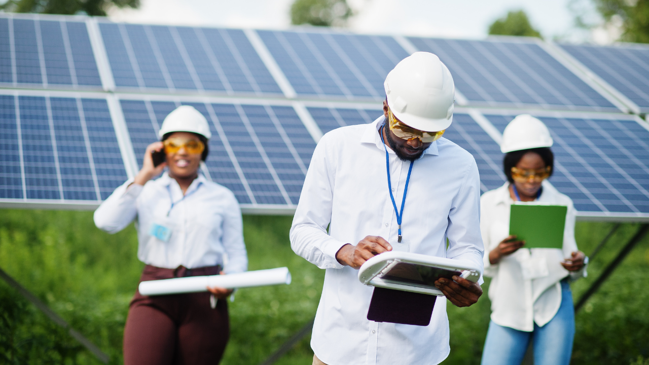 A team of engineers works independently in a solar farm, one of them on a call, another reviewing a tablet. Their focused activity hints at ongoing projects likely enabled by grants and subsidies for clean energy.