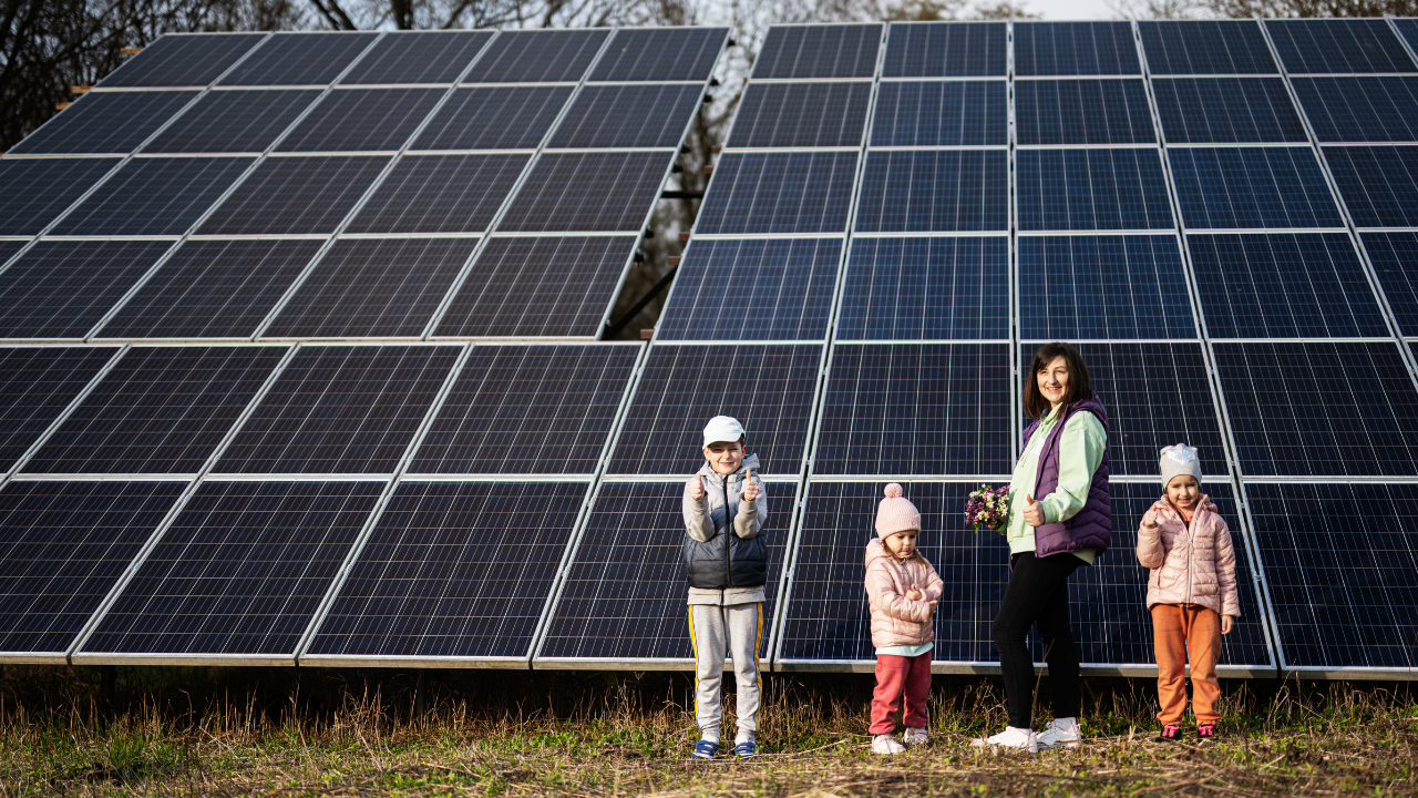 Una madre y cuatro niños posan frente a una estructura de paneles solares, sonriendo en un entorno rural. La imagen sugiere que el acceso a energía renovable es posible gracias a ayudas y subsidios gubernamentales.