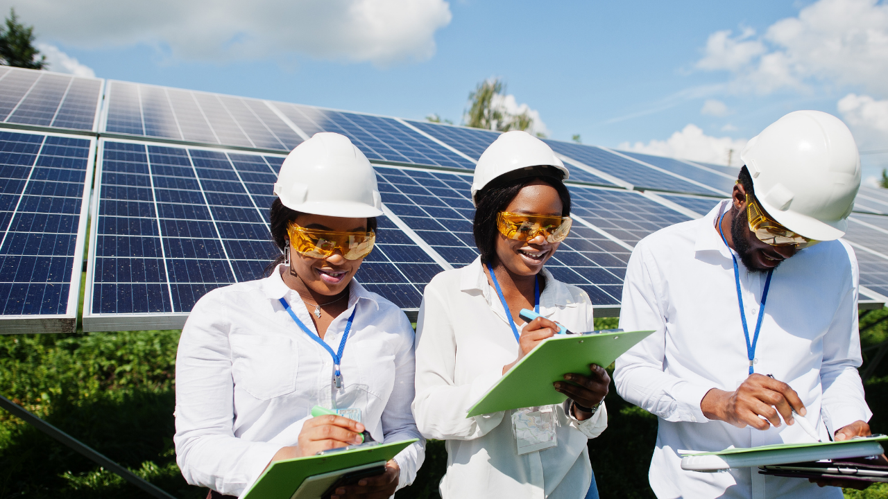 Tres técnicos con gafas protectoras toman notas frente a una hilera de paneles solares. Su trabajo meticuloso refleja cómo las ayudas y subsidios impulsan la profesionalización y expansión de la energía renovable en Uruguay.