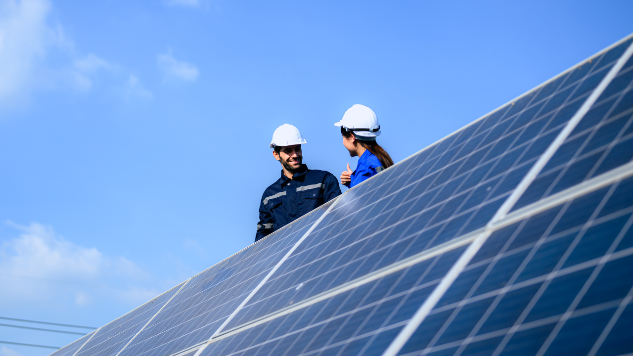 Two workers with helmets smile and converse beside tilted solar panels under a clear blue sky, illustrating a collaborative and optimistic American work culture when teams install solar panels.