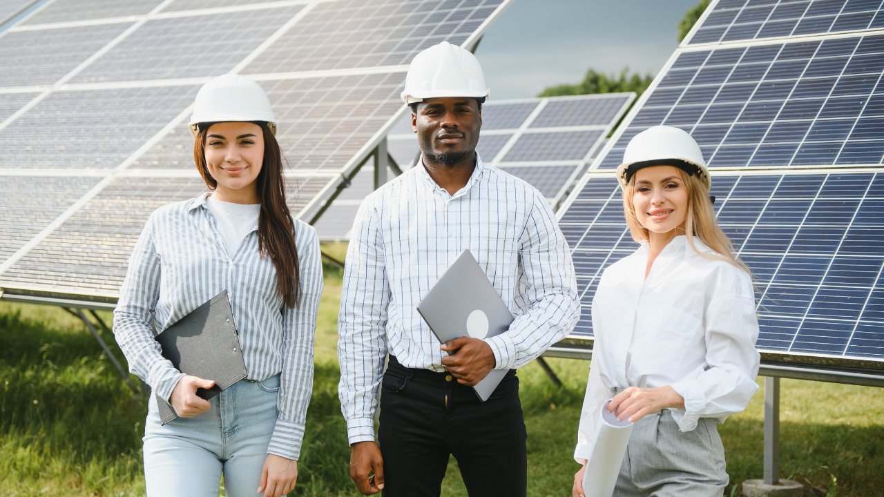 Tres personas posan con confianza frente a paneles solares. Llevan cascos y portan carpetas y dispositivos, representando el avance tecnológico en energía renovable mediante seguidores solares.