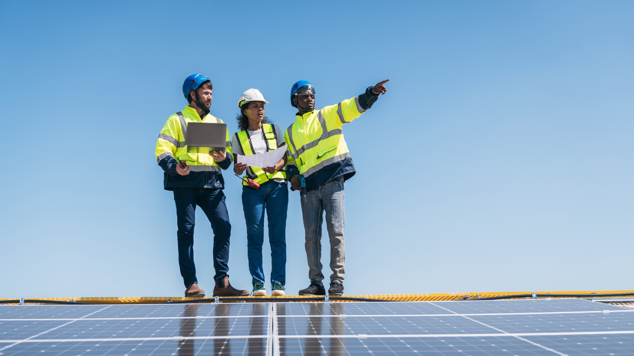 En una azotea con vista despejada, tres técnicos con chalecos reflectantes y cascos revisan planos mientras uno señala hacia el horizonte. La imagen transmite planificación y precisión en la instalación de seguidores solares.