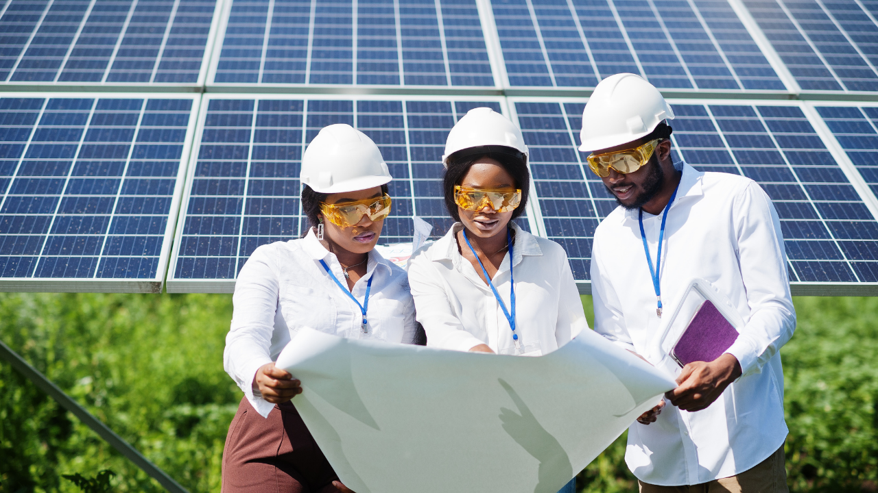 Three engineers wearing protective gear analyze a blueprint beneath a large solar array. The focus on planning and coordination reflects the installation and optimization of solar inverters in the UK’s green energy landscape.