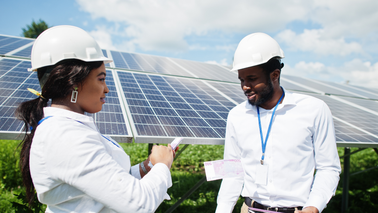 Dos trabajadores con cascos blancos inspeccionan billetes en mano, posiblemente discutiendo inversión o costos asociados. El fondo de paneles solares contextualiza el papel económico de los inversores solares en proyectos energéticos.