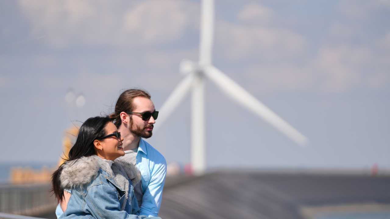 Una pareja disfruta del paisaje costero con una turbina eólica de fondo, mostrando la integración de lo sostenible en la vida cotidiana.