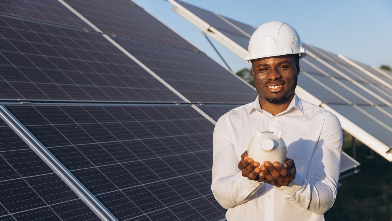 A smiling man in a hard hat holds a piggy bank in front of solar panels. His gesture symbolizes financial savings and sustainable choices, clearly linked to the benefits of portable solar panels.