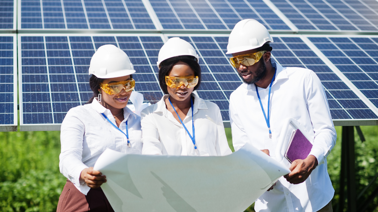 A team of three engineers examines a large blueprint in front of solar panels. Their focused collaboration highlights the strategic implementation of solar batteries for efficient energy storage.