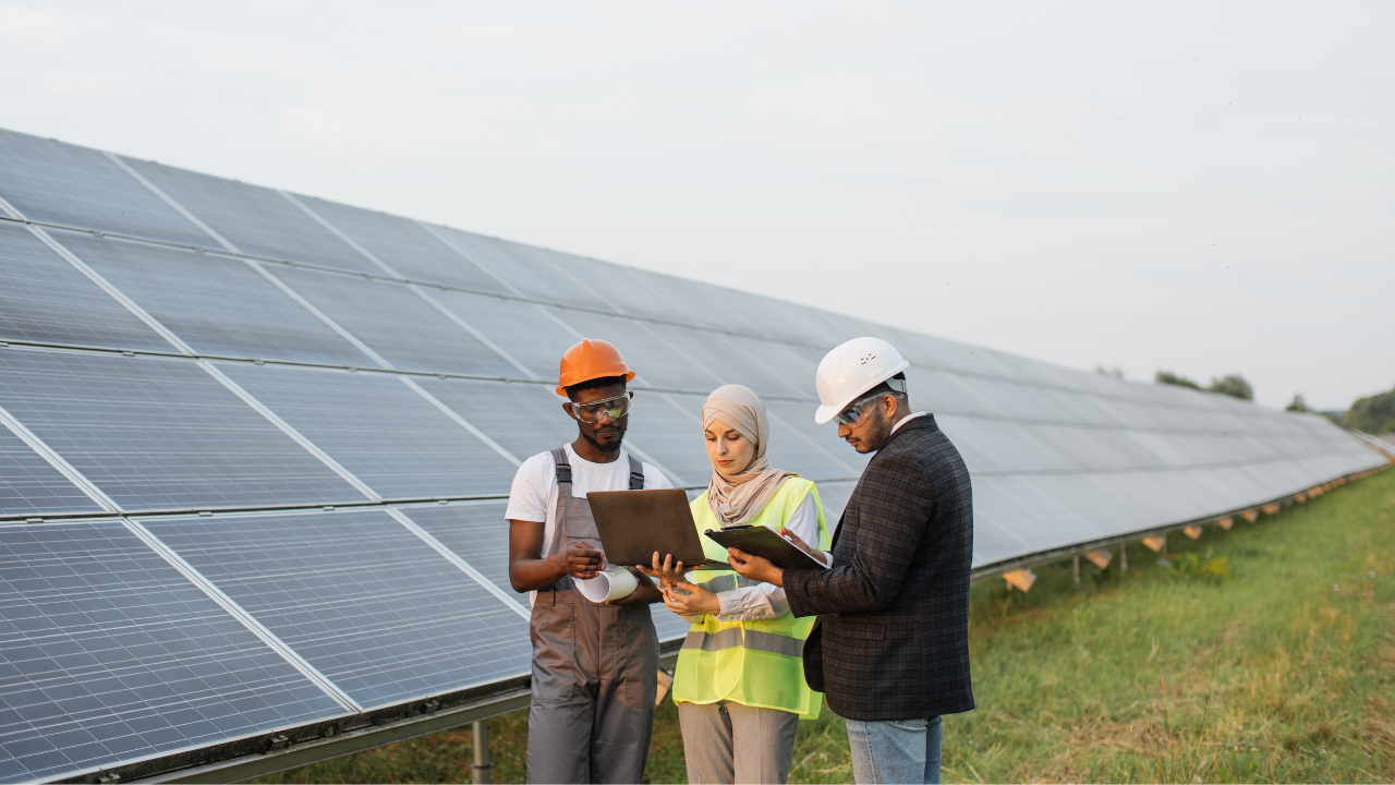 Un grupo diverso de profesionales revisa datos en laptops y carpetas frente a una extensa instalación solar. La escena resalta la planificación y análisis vinculados al uso de baterías solares para mejorar la autosuficiencia energética.