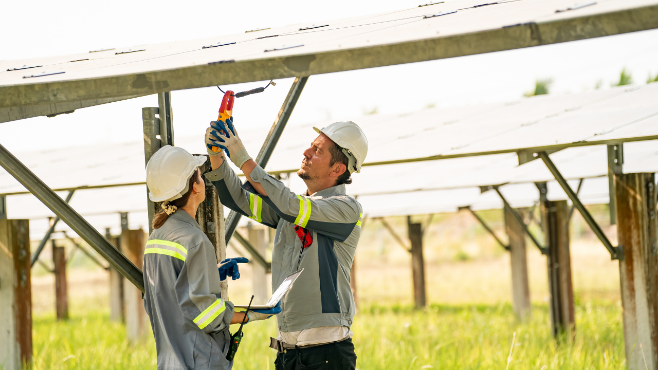 Dos técnicos realizan tareas de mantenimiento en la parte inferior de una instalación solar. Están midiendo componentes, lo que refleja una revisión técnica del sistema de paneles solares en funcionamiento.