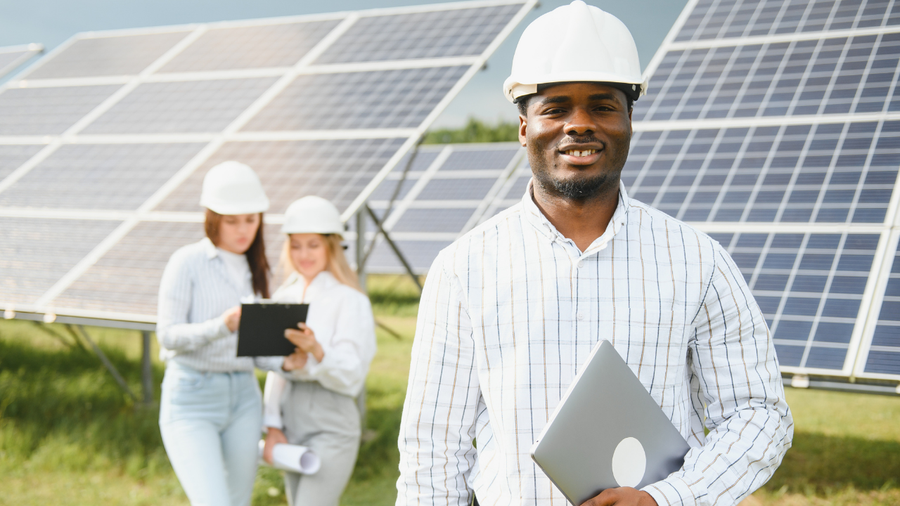 Un profesional sonríe a cámara con una laptop en mano, mientras dos colegas trabajan al fondo. El entorno solar sugiere gestión y supervisión operativa de un sistema de paneles solares.