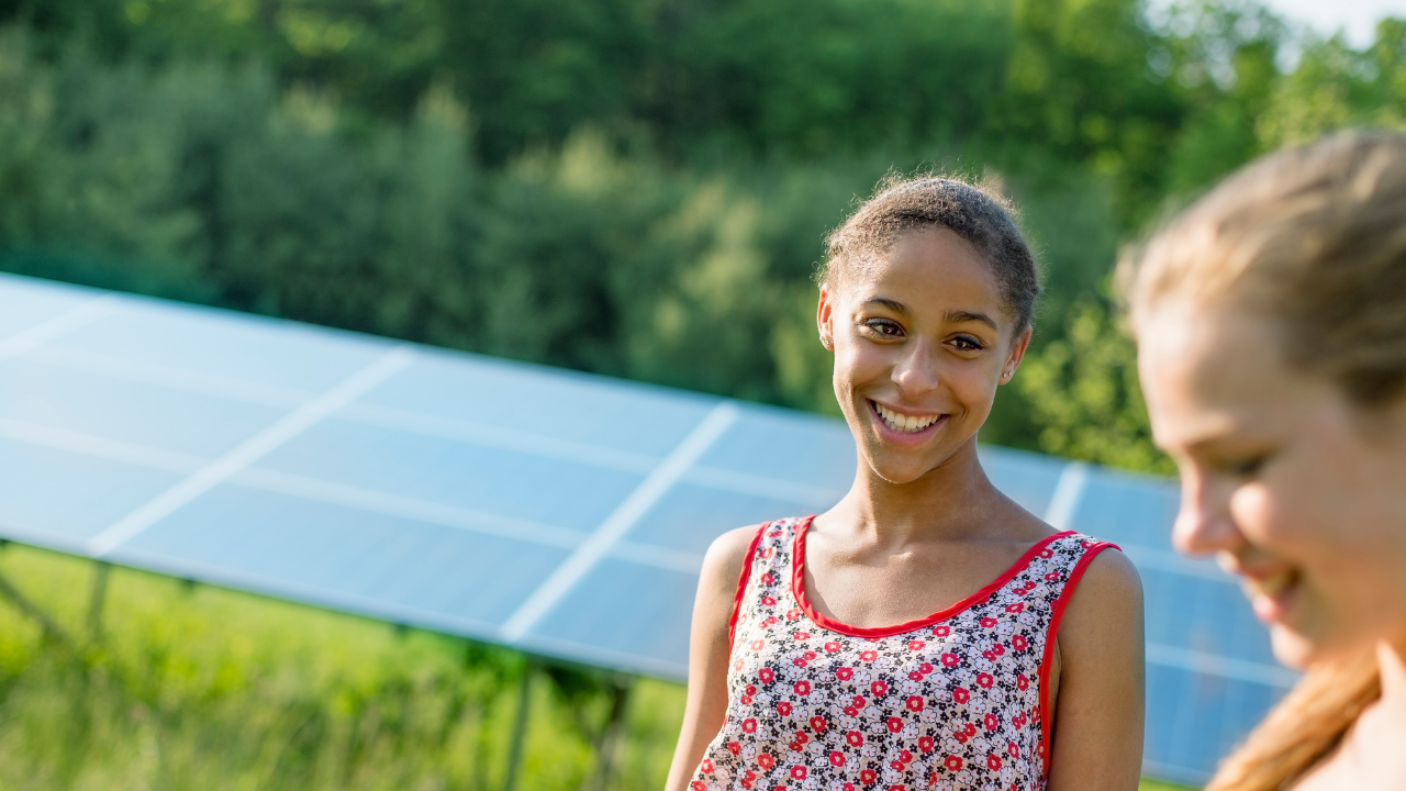 Una niña sonríe en un entorno natural con paneles solares al fondo. La escena representa el acceso comunitario a la energía fotovoltaica y plantea una visión comparativa con la energía térmica desde un enfoque cotidiano.