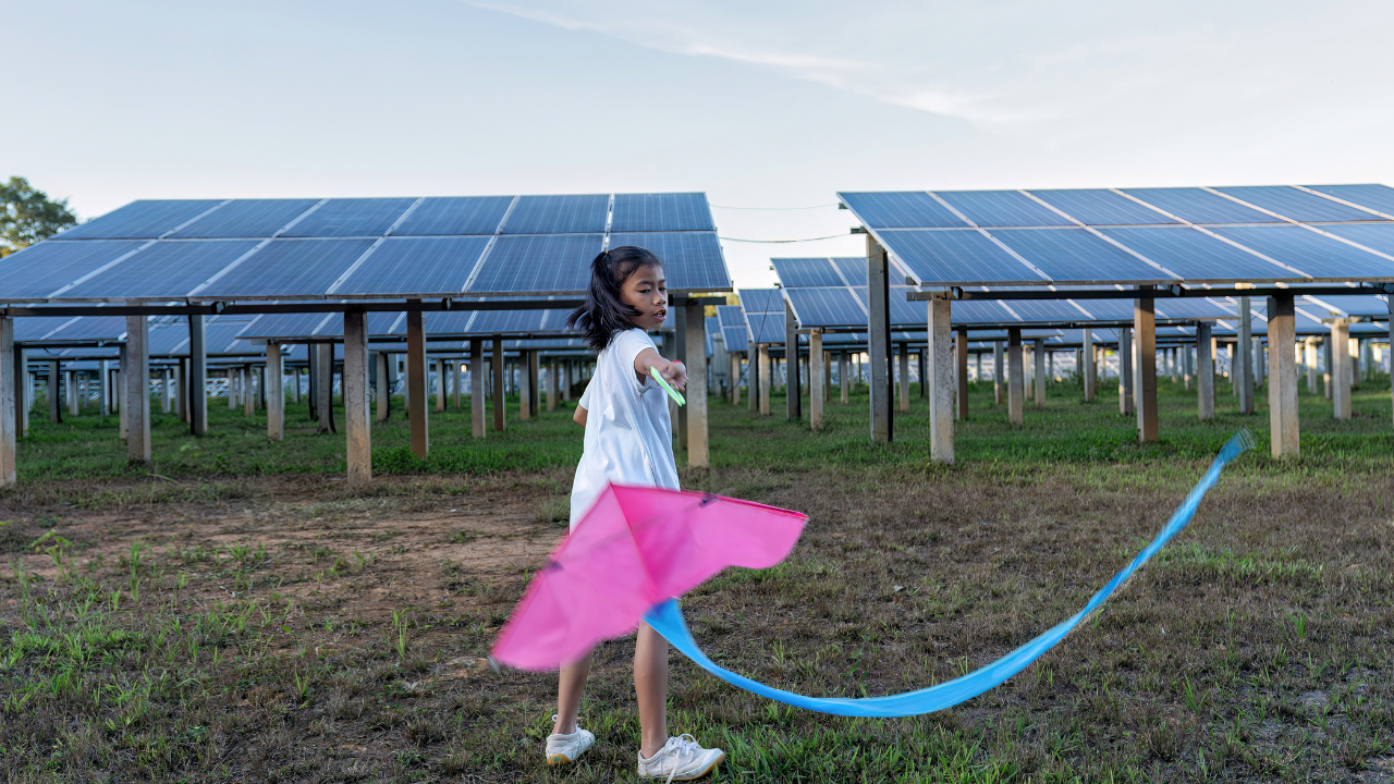 Una niña juega con una cometa entre paneles solares. La imagen ilustra la integración amigable de la energía fotovoltaica en la vida cotidiana, contrastando de forma visual su accesibilidad frente a opciones de energía térmica.
