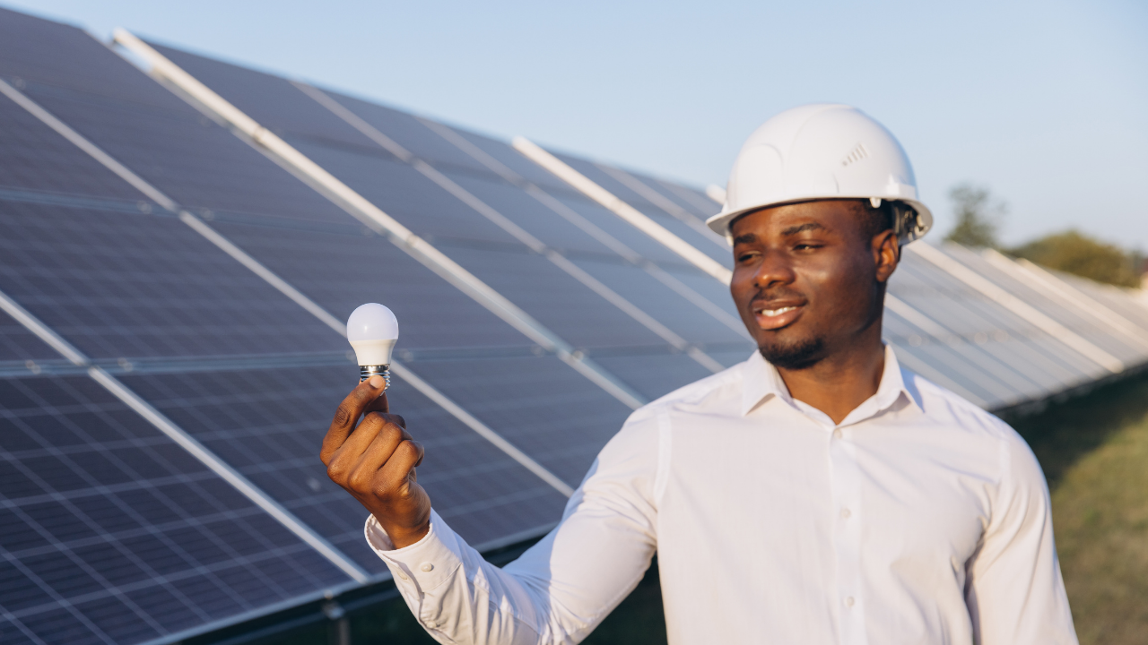 Un joven ingeniero observa una bombilla en su mano, con una amplia sonrisa, frente a un campo de paneles solares. La imagen comunica de forma simbólica el resultado de transformar la energía solar térmica en luz para el uso cotidiano.