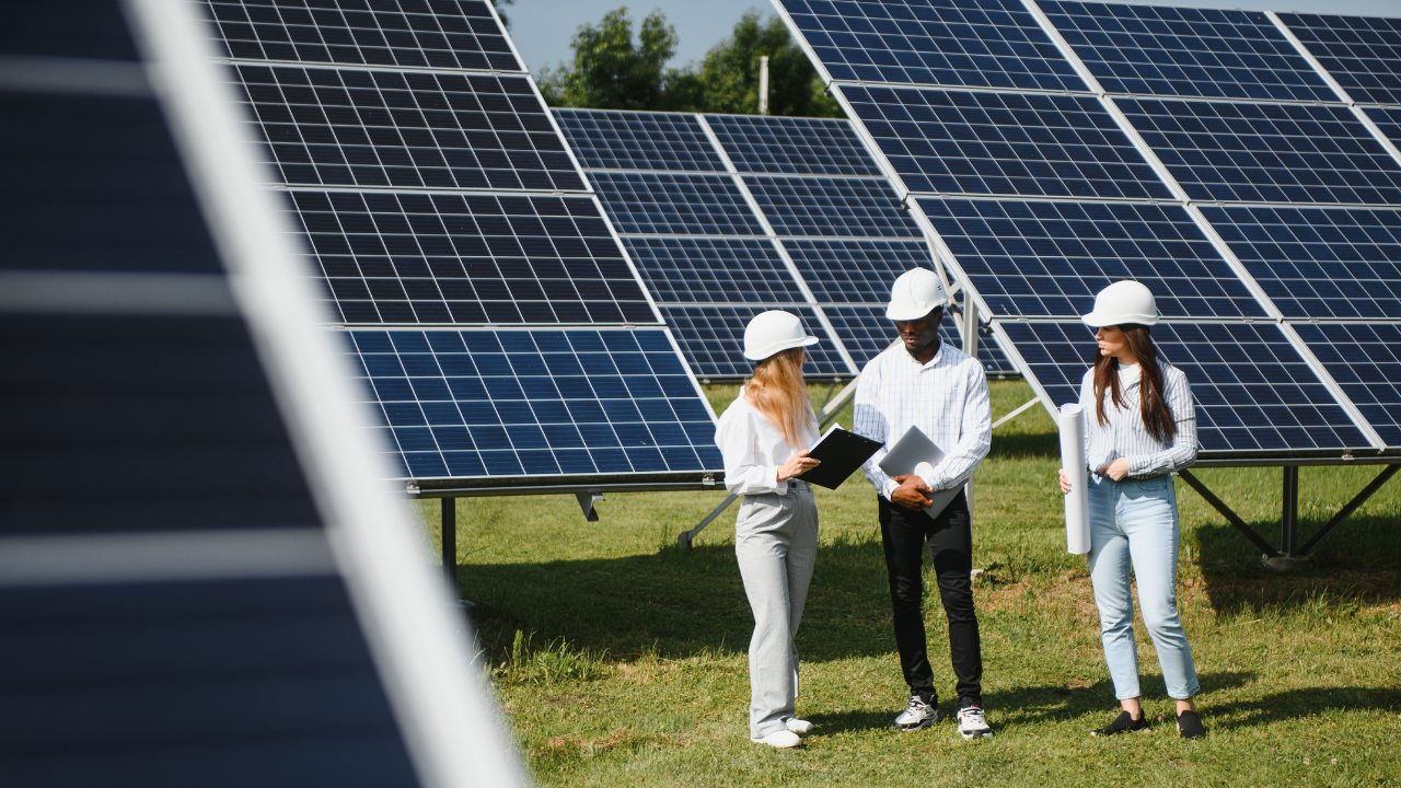 A group of engineers in white helmets stand among large solar panels, discussing a project while holding plans and tablets. The scene highlights a professional solar panel installer team working collaboratively under the British sun.