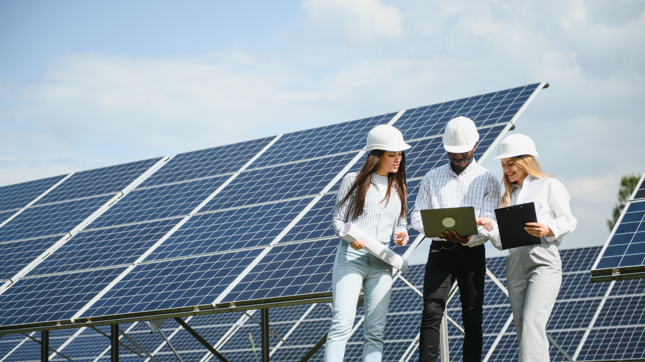 Tres personas, dos mujeres y un hombre, revisan información en laptops y planos frente a un campo de paneles solares. La escena refleja colaboración técnica e interdisciplinaria en el diseño y monitoreo de soluciones solares térmicas.