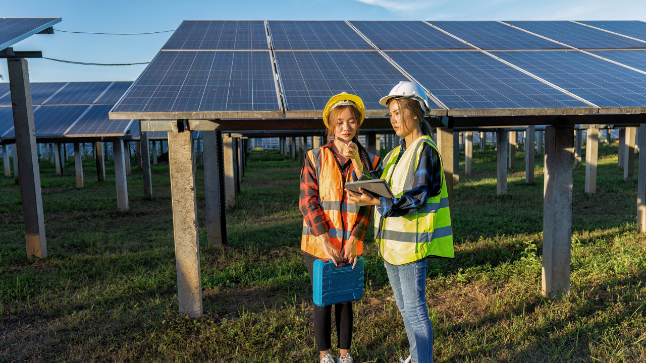 Two women in safety gear review data on a tablet while standing beneath solar panels, reflecting a hands-on approach to maintenance of solar panels in a field setting.