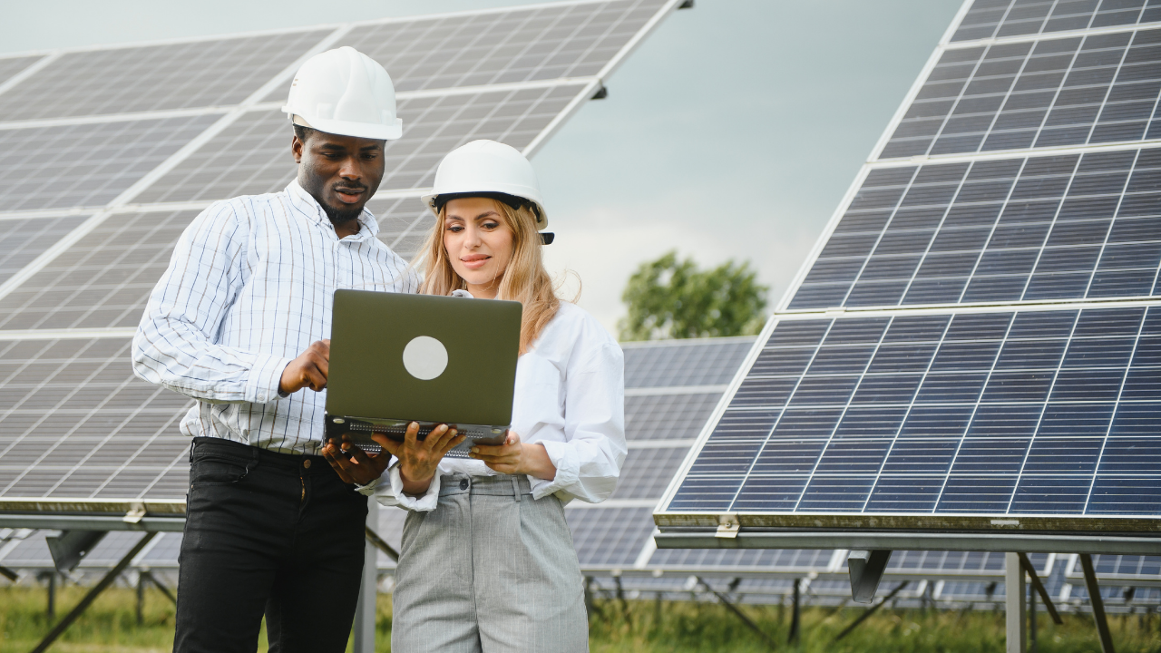 Un hombre y una mujer observan una computadora portátil frente a paneles solares, representando una colaboración técnica enfocada en el mantenimiento de paneles solares.