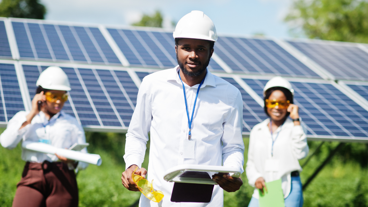 Un ingeniero al frente sostiene gafas protectoras y documentos mientras dos colegas al fondo interactúan, representando un equipo coordinado dedicado al mantenimiento de paneles solares.